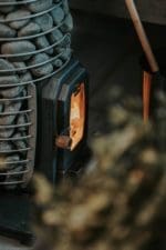 Image showcasing the HUUM HIVE Wood sauna heater, a modern embodiment of time-honored sauna heater construction practices. The heater's flame tamer, made of a robust 5 mm thick stainless steel, is highlighted, showcasing its exceptional durability. An available firebox extension is also depicted, illustrating the capability of heating the sauna from an adjacent room.